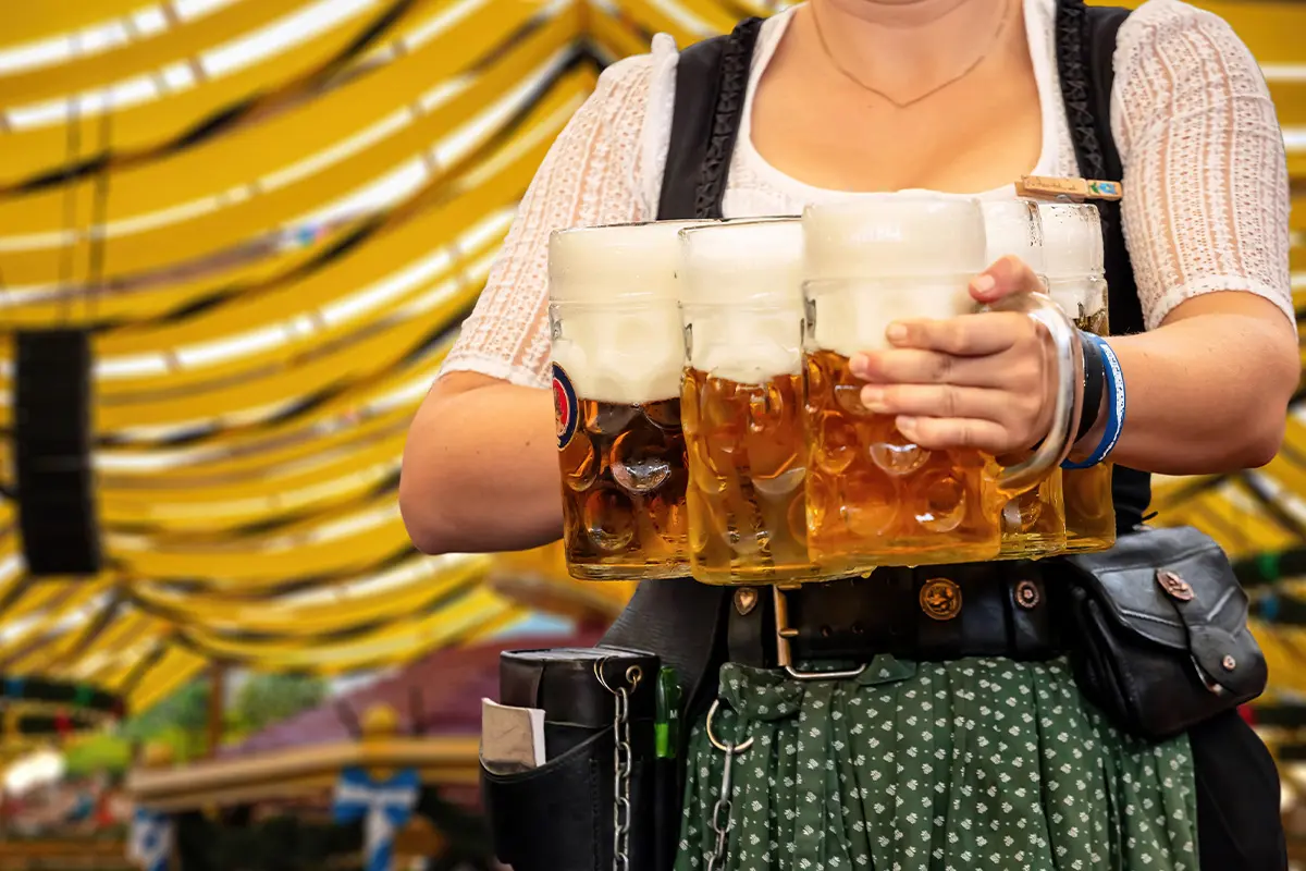 Waitress carrying large steins of frothy beer at a Bavarian festival, showcasing Oktoberfest drinks deals 2025