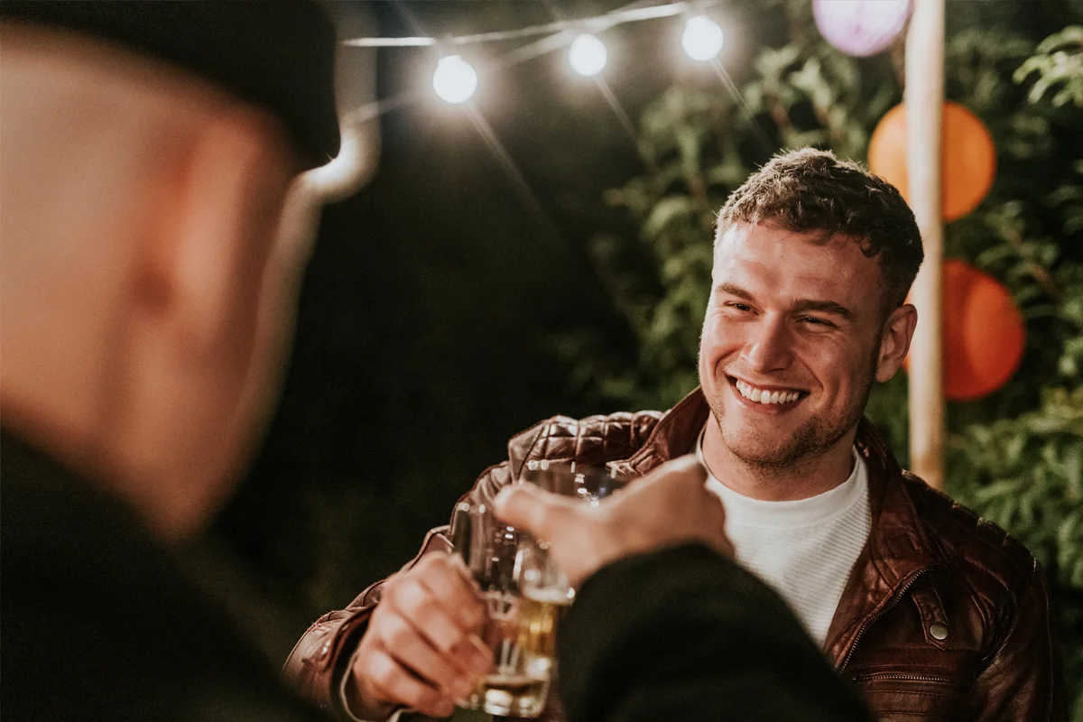 Two friends clinking glasses outdoors under warm string lights, with one smiling in a leather jacket.