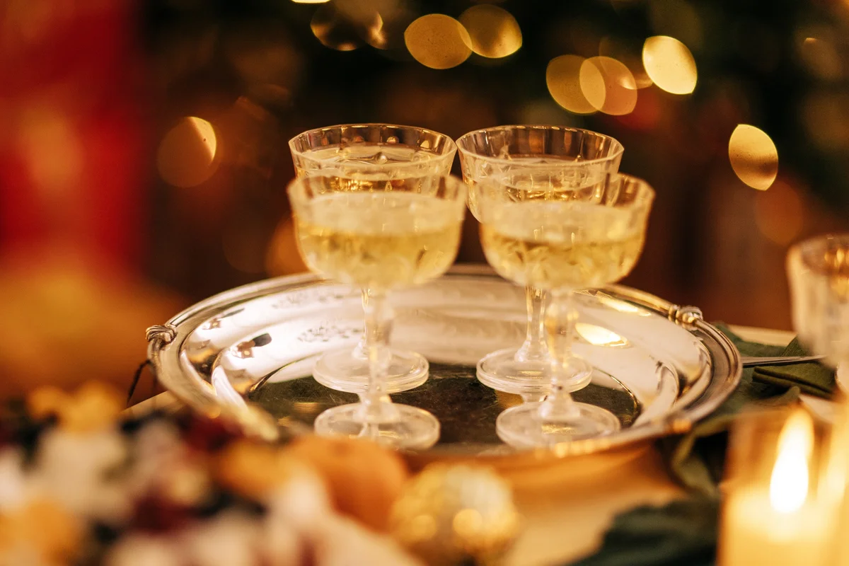 Four champagne coupes filled with sparkling wine on a polished silver tray, surrounded by soft golden festive lights.
