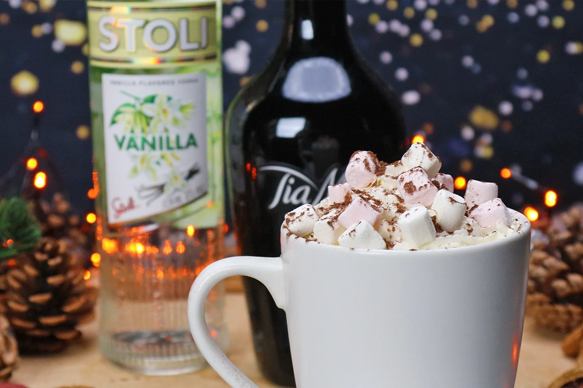 A mug of hot chocolate topped with marshmallows and cocoa, with vanilla vodka and coffee liqueur bottles behind it.