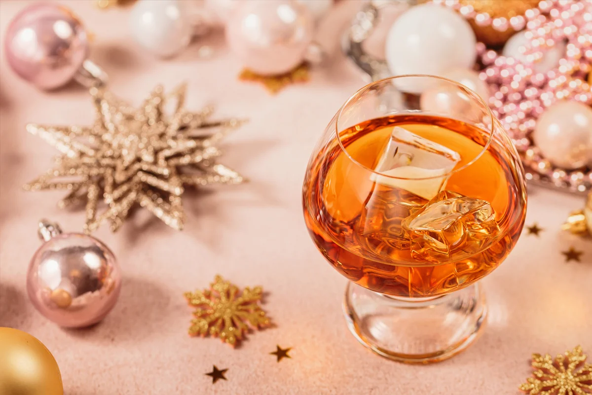 A glass of brandy with ice placed among sparkling Christmas baubles and golden festive decorations.