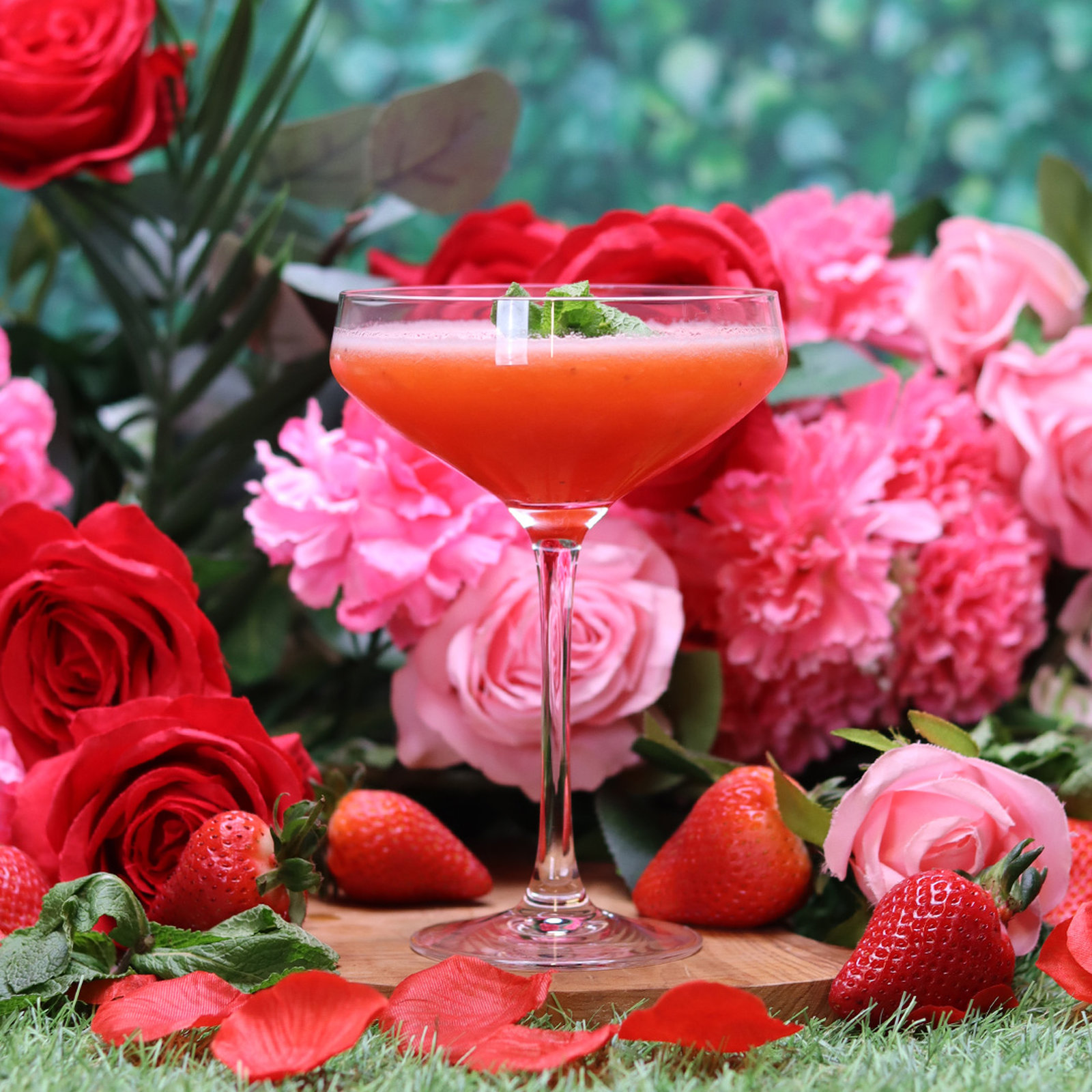 Stoli Strawberry Kiss cocktail in a coupe glass with mint garnish, surrounded by fresh strawberries, pink and red roses, featured as the cocktail of the month for bank holiday drinks deals near you at Wine Rack.